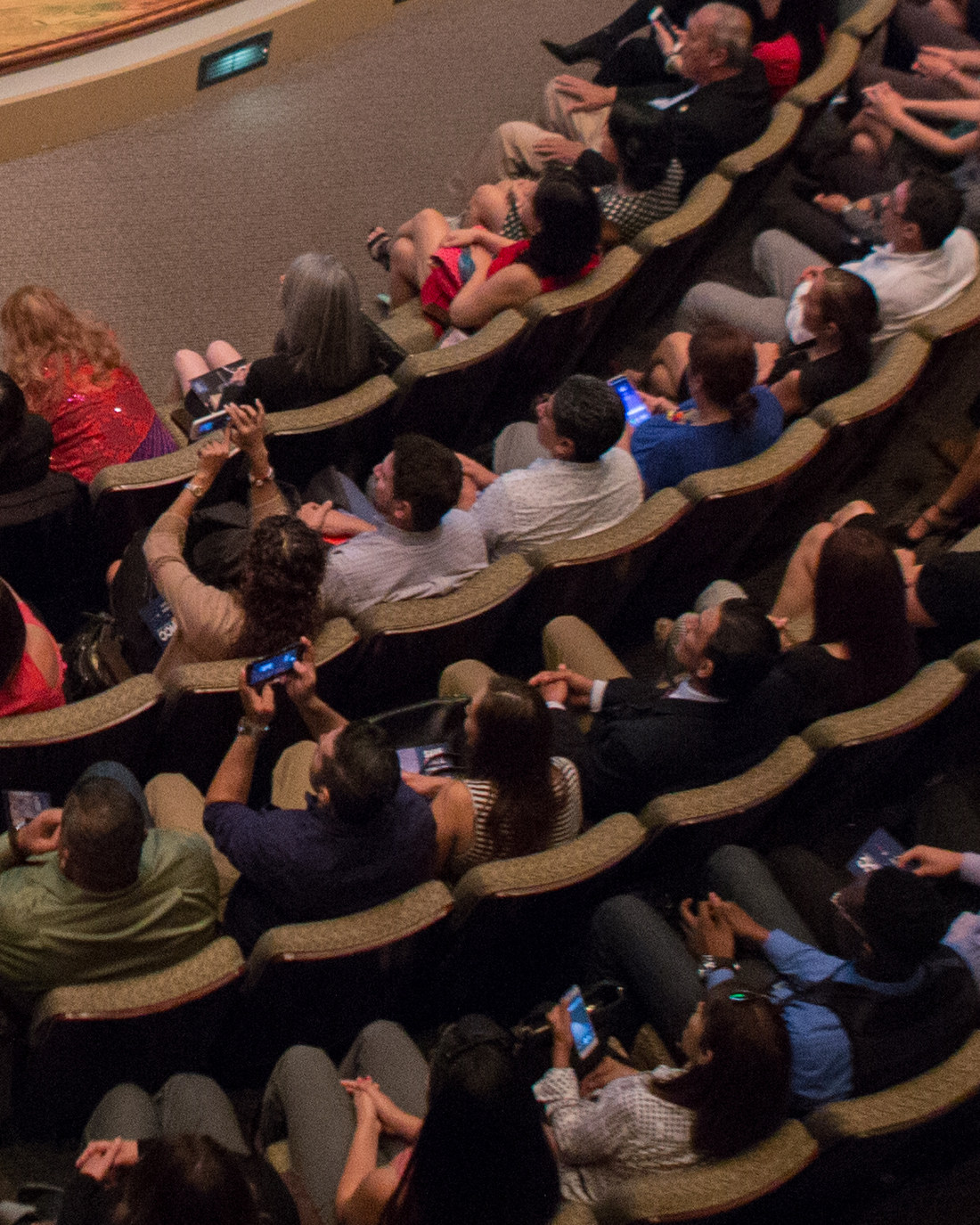 A full auditorium of attendees seated in tiered rows, watching and photographing a conversation on stage.